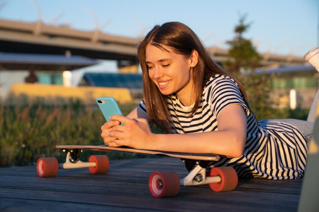 Happy Skateboarder Girl Hold Smartphone Messaging Use Mobile 5g Internet Outdoors For Social Media